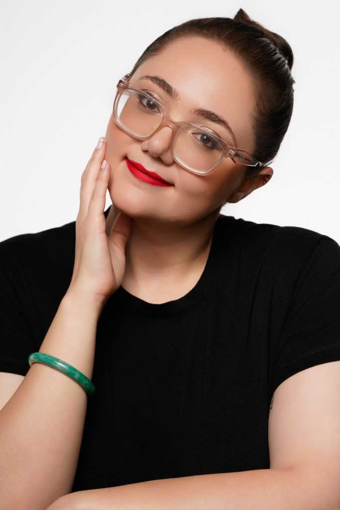 A studio portrait of a woman wearing clear-framed glasses and a black top, posing with her hand resting on her cheek against a plain white background.