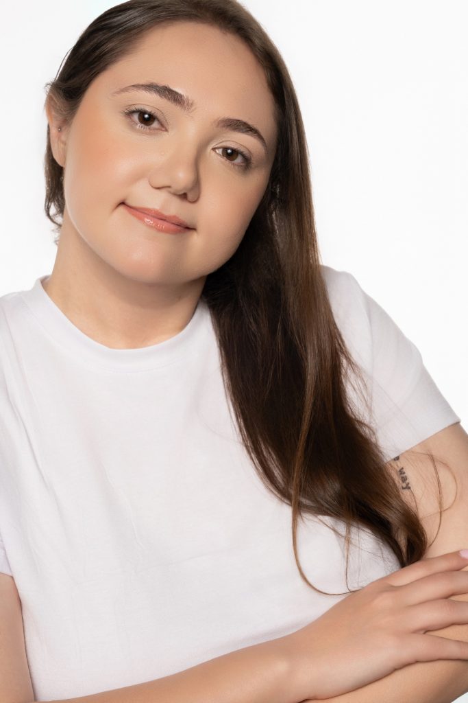 A softly lit studio portrait of a woman in a plain white shirt, looking directly at the camera against a white background.