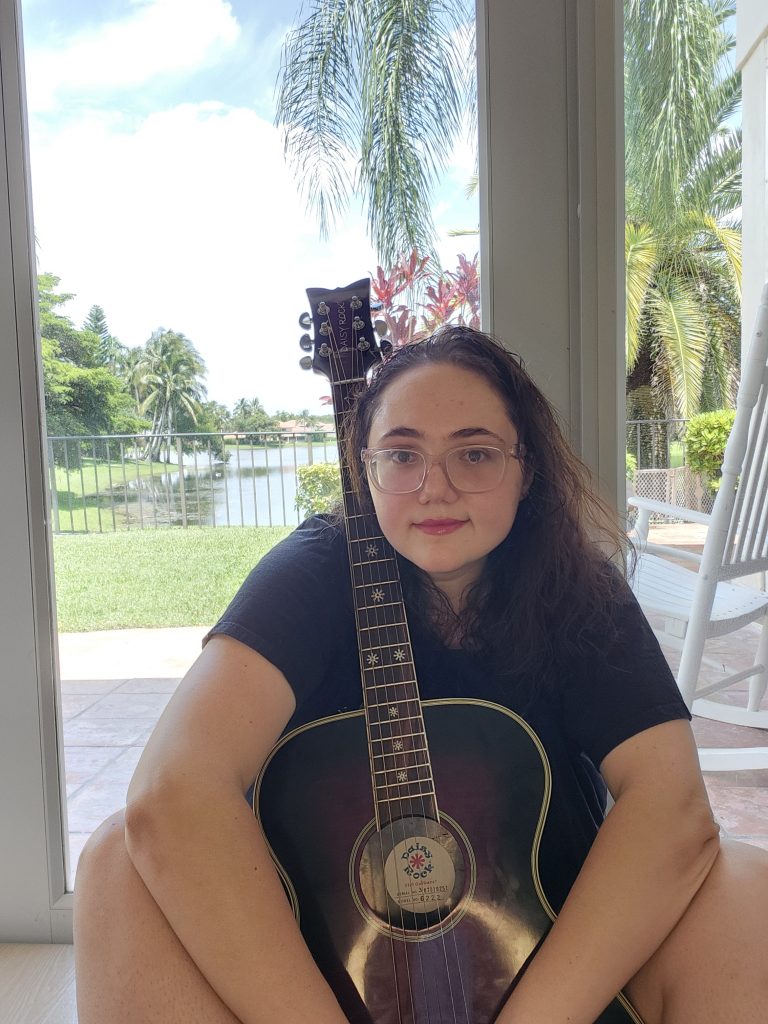 Lauren Grace Zemel sitting with acoustic guitar, natural light, lake and palm trees in background, South Florida