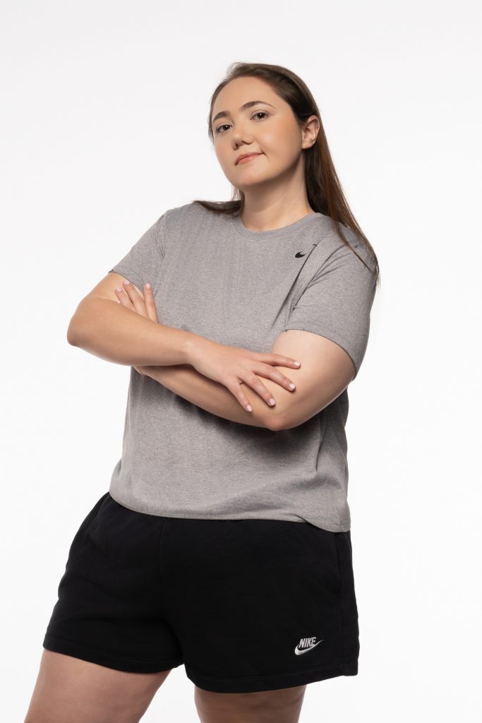 Lauren Grace Zemel smiling, arms crossed, grey Nike crop tee, black shorts, white studio background