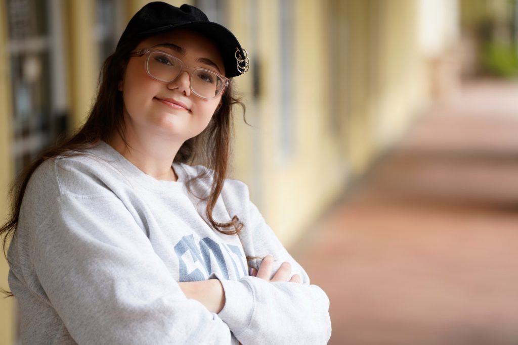 Lauren Grace Zemel smiling, arms crossed, Seoul South Korea sweatshirt, black cap, outdoor corridor background