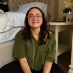 Lauren sitting cross-legged on her bedroom floor in a green striped shirt smiling at the camera