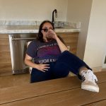 Lauren sitting on the floor in sunglasses and an I Swing Both Ways bisexual pride shirt holding a drink