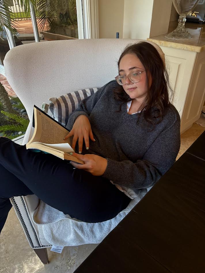 Lauren Grace Zemel curled up in a chair reading a book, wearing glasses and a gray sweater.