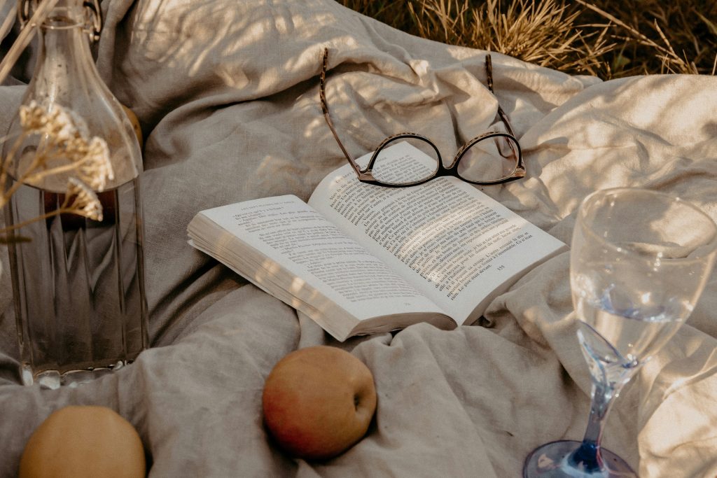An open book with reading glasses resting on it, surrounded by fruit and a wine glass on a linen blanket in golden light, representing the Fan Fiction category