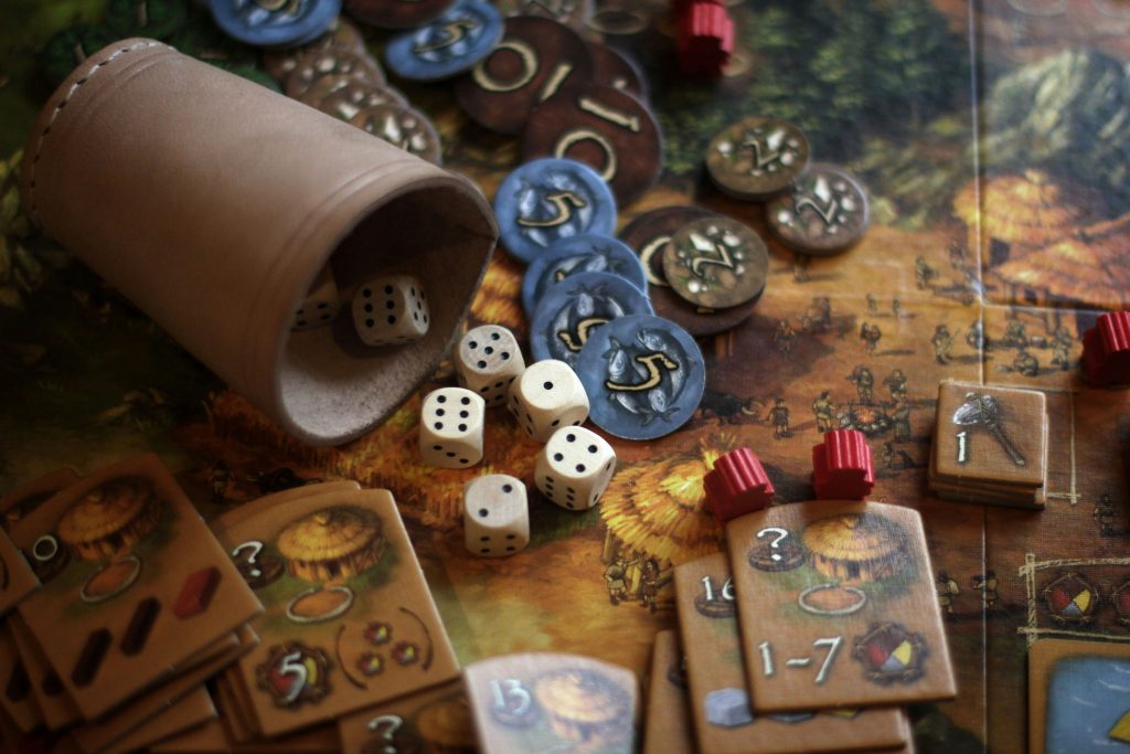 A close-up of tabletop game pieces including dice spilling from a leather cup, wooden tokens, and game cards, representing the Tabletops category