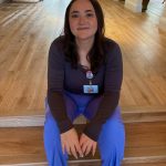 Lauren in blue scrubs and a dark top sitting on stairs with a badge