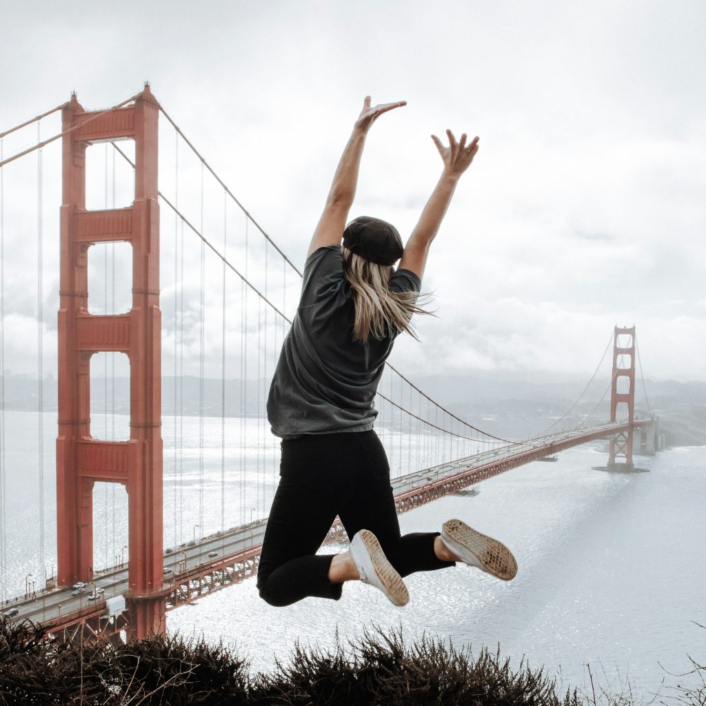 A woman jumping joyfully with arms raised in front of the Golden Gate Bridge, representing the Mind & Body category