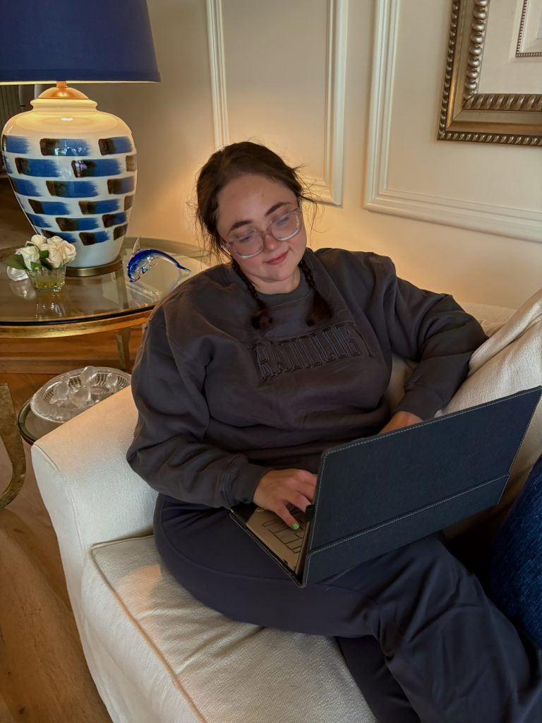 Radiologic technology student sitting on a couch, working on a laptop, wearing a gray sweatshirt.
